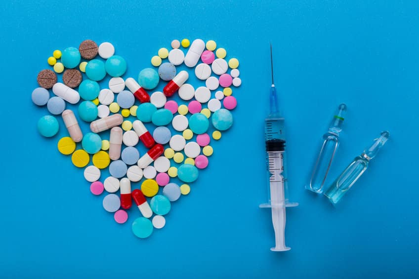 A lot of pills and syringe with injection on blue background with a heart shape as a symbol of medical treatment. Choice antibiotics against vaccine from Covid-19. Emergency help if a heart attack happened.