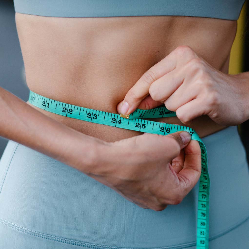 Close-up image of a person in athletic clothing measuring their waist with a tape measure to monitor weight loss progress.