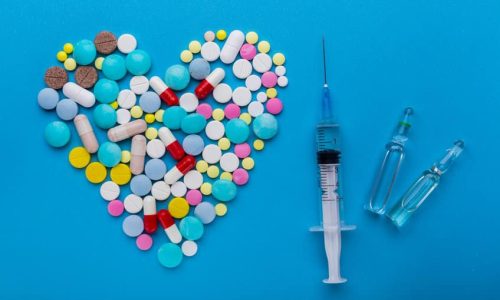 A lot of pills and syringe with injection on blue background with a heart shape as a symbol of medical treatment. Choice antibiotics against vaccine from Covid-19. Emergency help if a heart attack happened.